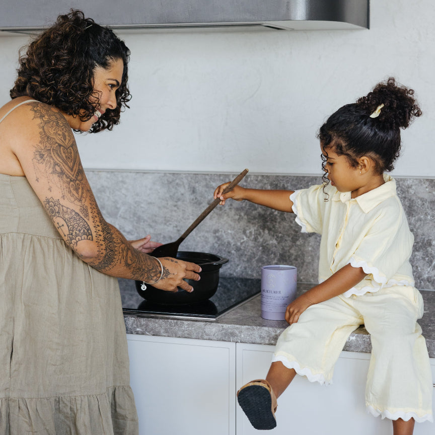 A woman with curly hair and tattoos stirs a pot in a modern kitchen while a young child in a cream colored ruffled outfit sits on the kitchen counter holding a wooden spoon