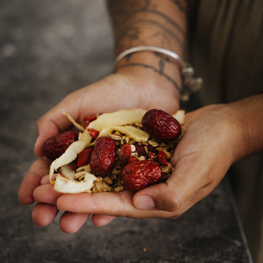A close up of a person's hands holding a small pile of herbal tea or loose leaf blend containing red dates, dried banana, and brown herbal pieces