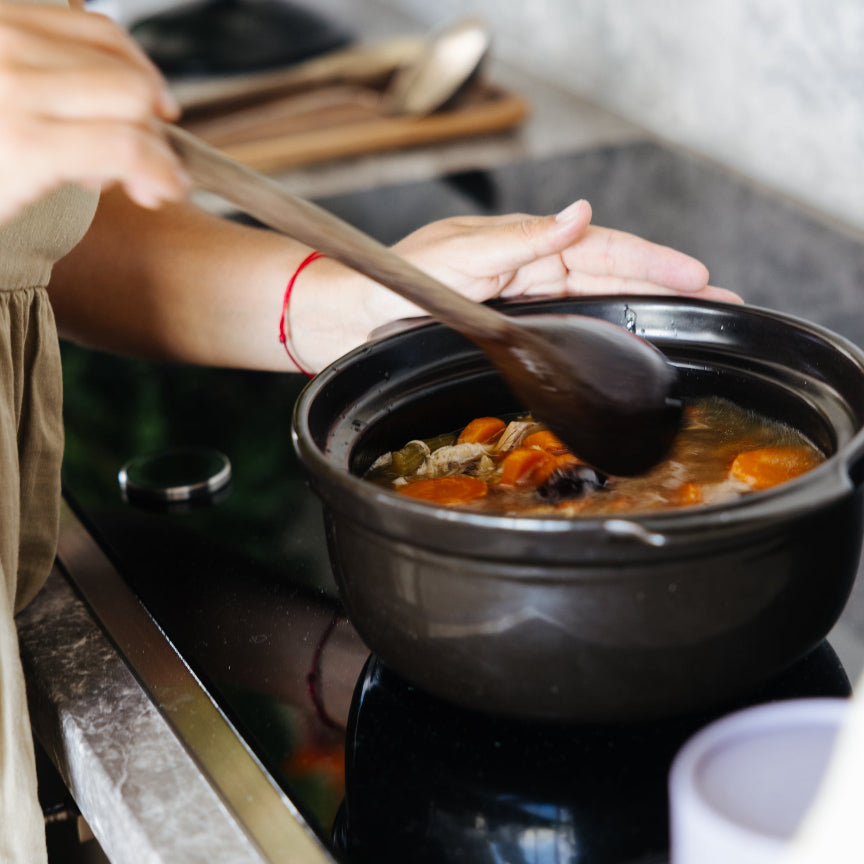 Segohealth_ Nurturer _ cooking potA person in a beige dress stirs a black clay pot filled with a vegetable soup on a black induction cooktop in a kitchen setting.