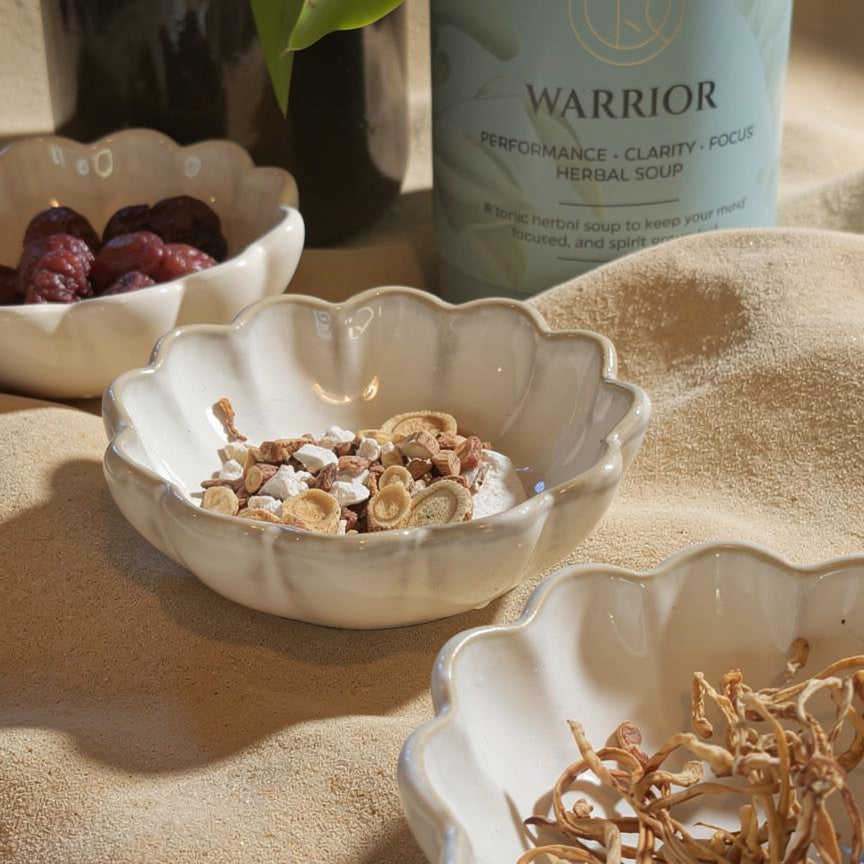 Segohelath  warrior herbal soup is styled on a beige fabric surface with small white scalloped bowls containing dried herbs and red dates in the foreground